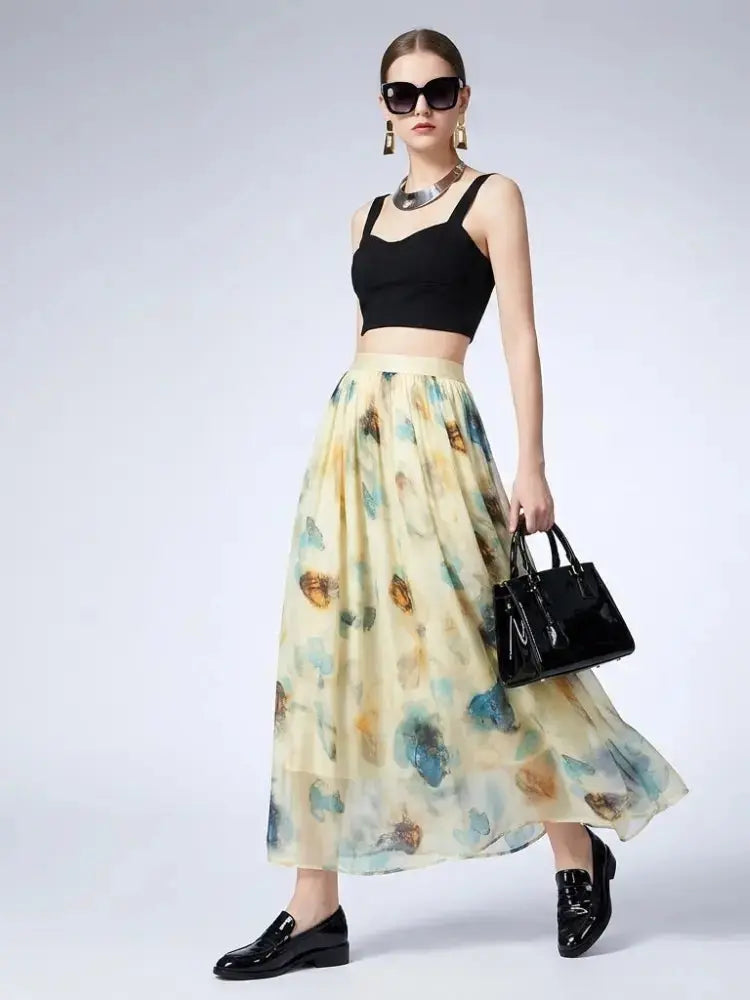 A model wearing a black crop top and a long, flowing cream skirt with a blue and brown watercolor floral print. She is accessorized with black sunglasses, a silver statement necklace, gold earrings, a black handbag, and black loafers, set against a plain light gray studio background.