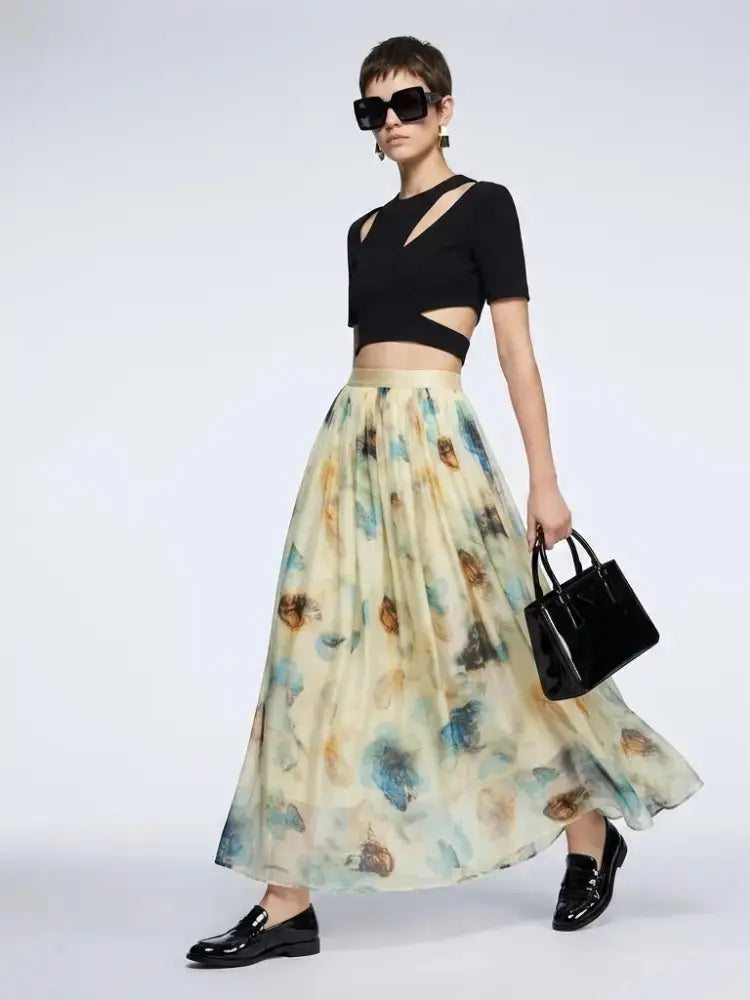 A model wearing a black short-sleeve crop top with cutouts, a long cream-colored skirt with a blue and brown floral print, black sunglasses, and black loafers, holding a black handbag against a light gray studio background.