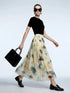 A model wearing a black short-sleeve top and a flowing cream-colored midi skirt with a blue and brown floral print, holding a black handbag and wearing black loafers against a light gray studio background.