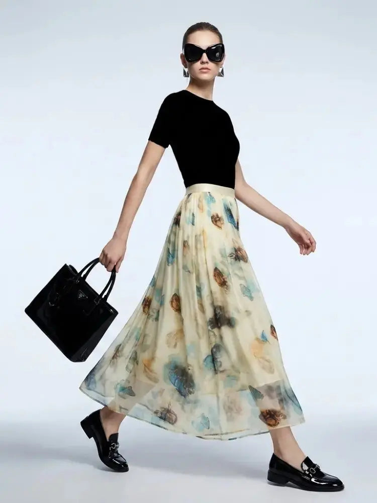 A model wearing a black short-sleeve top and a flowing cream-colored midi skirt with a blue and brown floral print, holding a black handbag and wearing black loafers against a light gray studio background.