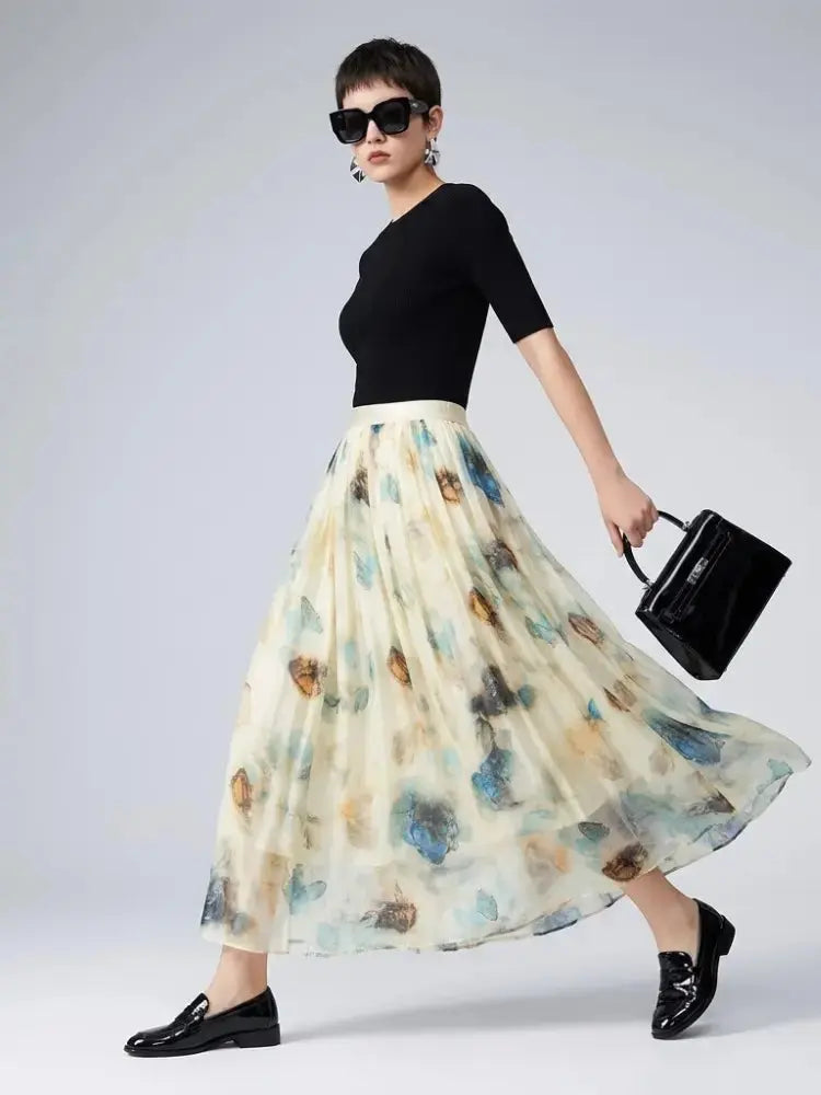 A woman models a high-waisted, full-length pleated midi skirt with a watercolor floral print in blue, brown, and beige on a cream base, paired with a black top, black loafers, sunglasses, and a black handbag against a light gray studio background.