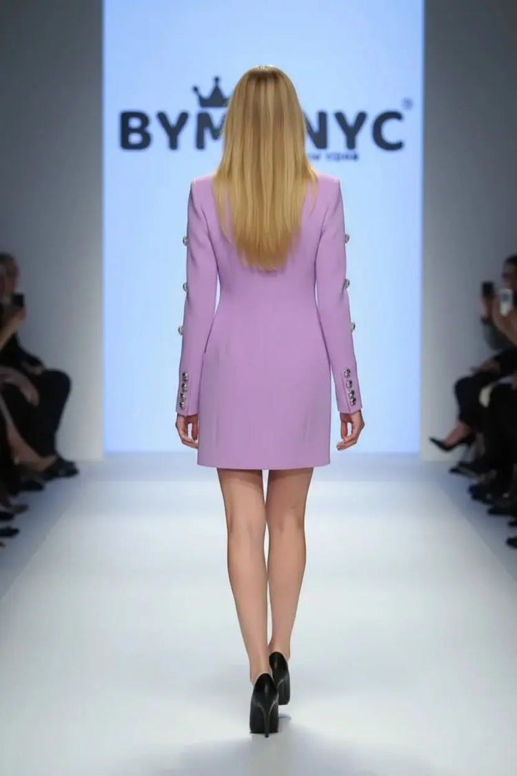 A model walks the runway wearing a lavender purple blazer-style mini dress with structured shoulders and decorative buttons. The background features a large BYM NYC logo on a white screen, with audience members visible on both sides of the catwalk.
