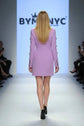 A model walks the runway wearing a lavender purple blazer-style mini dress with structured shoulders and decorative buttons. The background features a large BYM NYC logo on a white screen, with audience members visible on both sides of the catwalk.