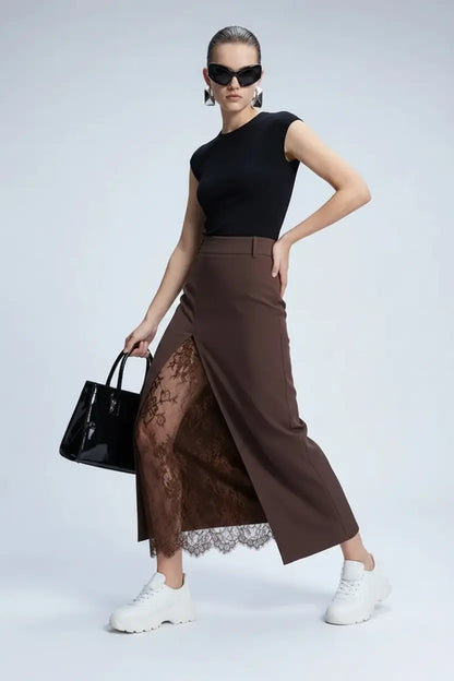 A model poses in a studio wearing a black sleeveless top, a brown high-waisted midi skirt with a sheer lace overlay, black socks, and patent leather shoes, while holding a black handbag.