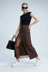 A model poses in a studio wearing a black sleeveless top, a brown high-waisted midi skirt with a sheer lace overlay, black socks, and patent leather shoes, while holding a black handbag.