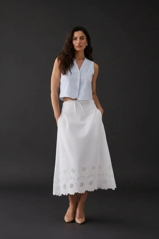 A woman stands against a plain light gray studio background wearing a sleeveless light blue cropped top and a long white skirt with embroidered detailing near the hem, paired with white flats.