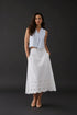 A woman stands against a plain light gray studio background wearing a sleeveless light blue cropped top and a long white skirt with embroidered detailing near the hem, paired with white flats.