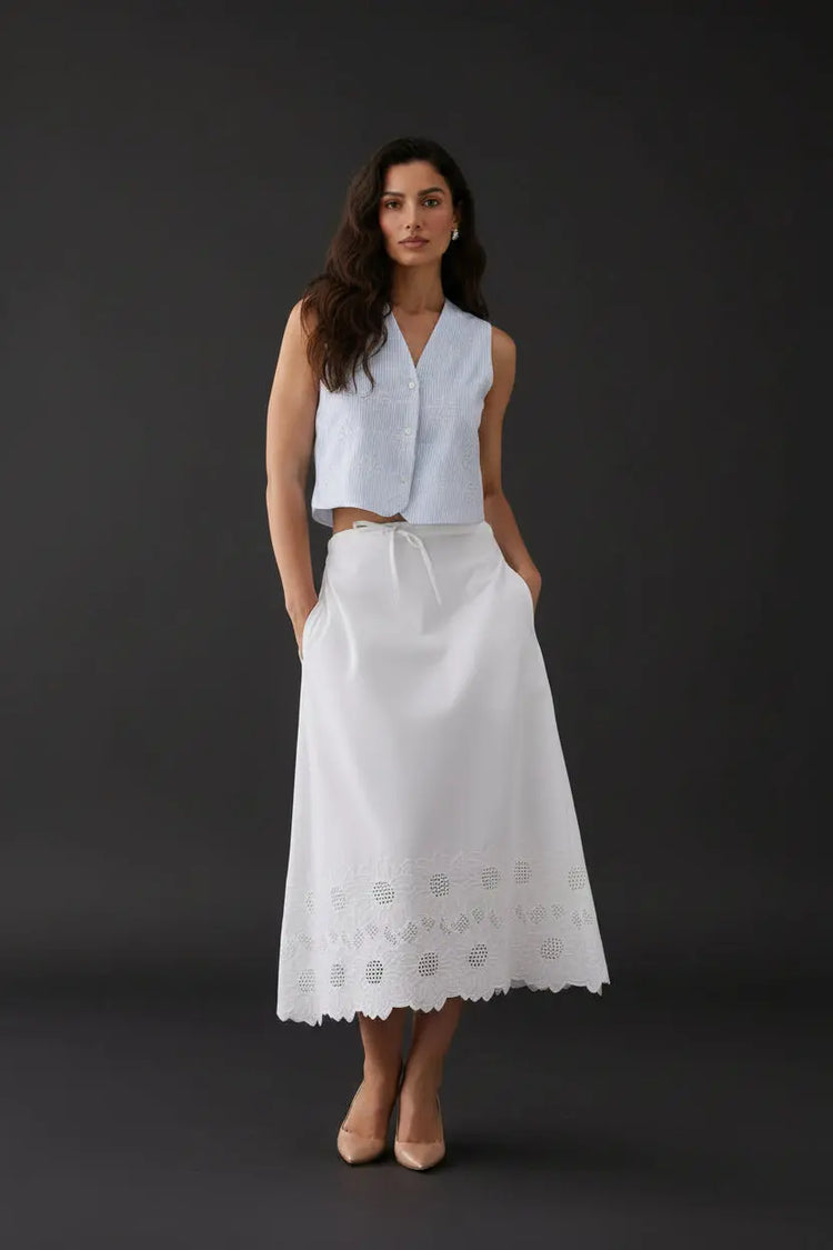 A woman stands against a plain light gray studio background wearing a sleeveless light blue cropped top and a long white skirt with embroidered detailing near the hem, paired with white flats.