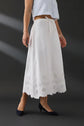 A model wearing a white midi skirt with a scalloped eyelet lace hem, styled with a light blue cropped top and white ballet flats against a plain light gray studio background.